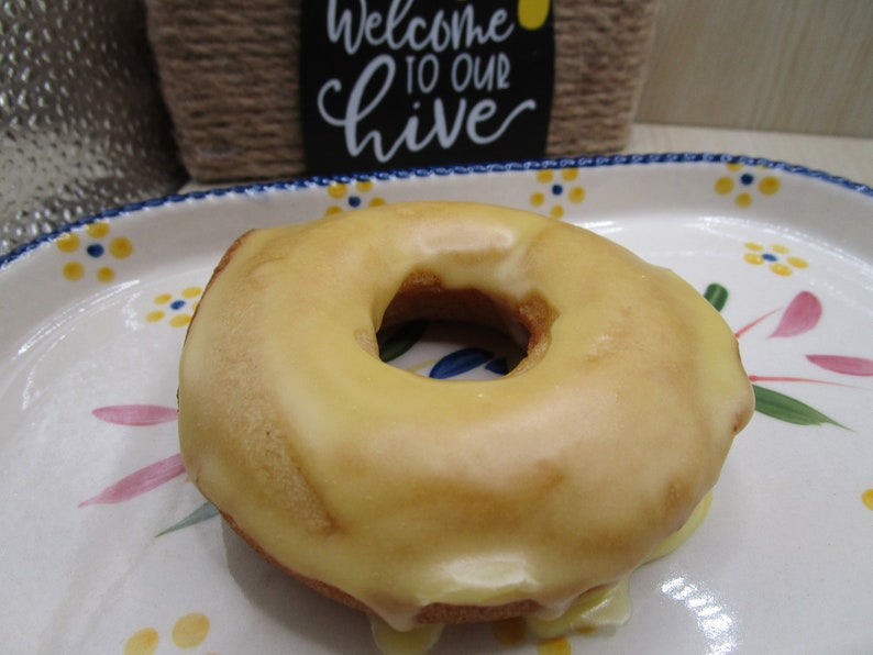 Puede incluir: Un d&oacute;nut redondo de color marr&oacute;n dorado con un glaseado suave de color amarillo p&aacute;lido, sobre un plato ovalado de cer&aacute;mica blanca decorado con peque&ntilde;os motivos florales azules y amarillos y p&eacute;talos rosas. Se ve un cartel con Welcome to our hive.