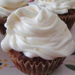 May include: A brown cupcake topped with a generous swirl of thick, white frosting. The cupcake is encased in a pleated, light brown paper liner. The frosting is applied in a smooth, continuous motion, creating soft ridges and peaks across the top.