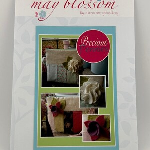 May include: A craft pattern titled "May Blossom" by Simone Gooding. The cover features a collage of images showcasing a beige pouch with floral embellishments. The text "Precious Pennies" is in a red circle. The design includes a light blue and green border.