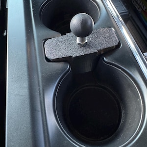 May include: A black textured car cup holder mount with a black ball and socket joint. The mount is designed to fit into a car's existing cup holders. The ball joint allows for adjustable positioning of devices.