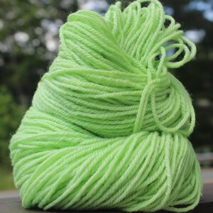 May include: A close-up of a ball of bright green yarn. The yarn is tightly wound, with loose strands visible at the top. The background is blurred, with hints of green foliage.