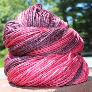 May include: A skein of yarn in a pink and purple ombre colorway. The yarn is wound into a ball shape and is ready to be used for knitting or crocheting.