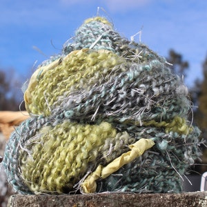 May include: A ball of yarn in shades of green, blue, and gray. The yarn is fluffy and textured, with a loose, hand-spun look.