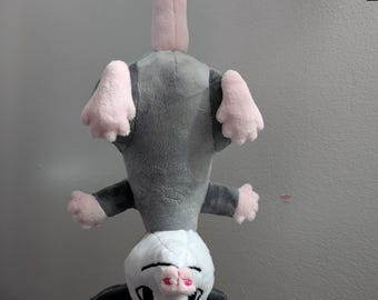 May include: A plush opossum toy, hanging upside down. The opossum has a gray body, pink paws and ears, and a white face with black eyes and a pink nose. The toy is suspended from a black metal bar.