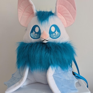 Può includere: Un giocattolo a forma di pipistrello di peluche con corpo bianco, dettagli blu e grandi orecchie foderate di rosa. Il pipistrello ha un colletto blu soffice e occhi ricamati. È seduto su un contenitore di plastica bianco.