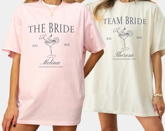 May include: Two pink and white t-shirts with a cocktail graphic and the text "The Bride" and "Team Bride". The shirts also include the text "Juli 2025" and the names "Melina" and "Theresa".