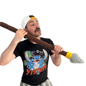 Puede incluir: Una persona con una camiseta negra con un personaje de dibujos animados azul, una gorra de béisbol blanca con visera amarilla y pantalones cortos de estampado gris y blanco. La persona sostiene una lanza de crochet marrón y gris con una punta amarilla.