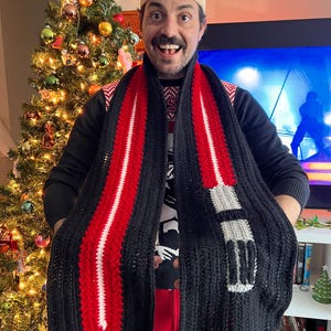 Könnte beinhalten: Ein schwarz-roter gehäkelter Schal mit einem Lichtschwert-Design. Der Schal hat ein rot-weißes Klingenmuster mit einem grau-weißen Griff. Der Schal wird über einem schwarzen Pullover mit einem rot-weißen Muster getragen.
