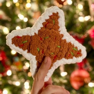 Puede incluir: Un adorno de estrella de jengibre de color marrón tejido a crochet con glaseado blanco y chispas verdes y rojas. El adorno se sostiene en una mano contra un fondo borroso de un árbol de Navidad con luces.