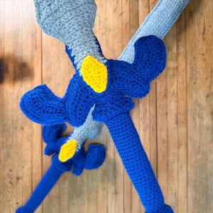 Peut inclure: Deux épées faites à la main au crochet. Les épées ont une lame et une poignée bleues avec des accents jaunes. La garde et la poignée sont bleues, et la lame est bleu clair et grise. Les épées sont posées sur une surface en bois.