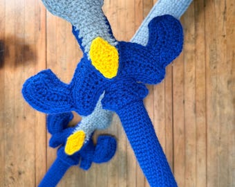 May include: Two handmade crochet swords. The swords feature a blue blade and handle with yellow accents. The hilt and handle are blue, and the blade is a lighter blue and gray. The swords are lying on a wooden surface.
