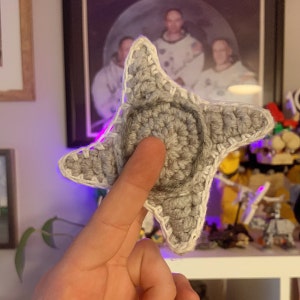 May include: A grey, crocheted star-shaped item with a white border. The star has a circular centre and pointed edges. The item is handmade and appears to be soft. The background includes a framed photograph and other decorative items.