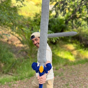May include: A handmade crochet sword featuring a long grey blade, a blue crossguard with a central yellow accent, and a blue handle. The sword is crafted with visible yarn stitches, showcasing a textured, soft construction throughout the entire piece.