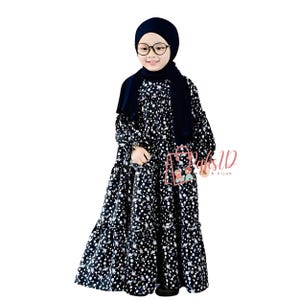 May include: A young person wearing a black and white floral dress with a navy blue hijab. The dress has a ruffled hem and long sleeves. The person is wearing glasses and has a neutral expression.