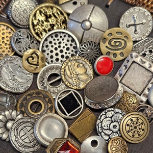 May include: A collection of vintage metal buttons in various shapes, sizes, and designs. The buttons are a mix of silver, gold, and brass tones, with some featuring intricate patterns and others with simple geometric shapes. There are also a few red glass buttons.