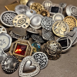 May include: A collection of vintage-style metal buttons in various shapes, sizes, and designs. The buttons feature a mix of silver, gold, and black finishes, with intricate details and patterns. Some buttons have a raised design, while others are flat.