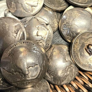 May include: A collection of silver-toned metal buttons crafted from vintage United States Buffalo nickel coins. Each button features the profile of a Native American or the image of an American bison, with a metal shank attached to the reverse side.