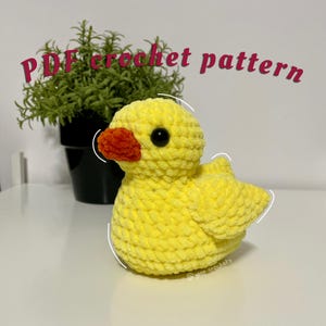 Może przedstawiać: Żółta dziana kaczka z czerwonym dziobem i czarnymi oczami. Kaczka siedzi na białym tle. Tekst "PDF crochet pattern" jest napisany na różowo nad kaczką.