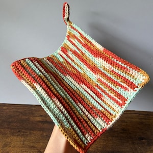 May include: A crocheted pot holder with a striped pattern in shades of orange, green, and brown. The pot holder has a loop at the top for hanging.