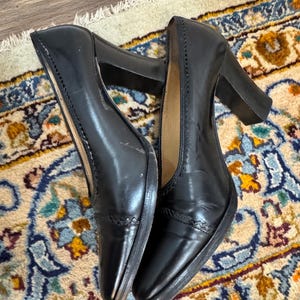 May include: A pair of black leather pumps with block heels. The shoes have a pointed toe and decorative stitching along the top edge. The heels are approximately 3 inches high. The shoes are displayed on a patterned rug.