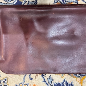 May include: A burgundy leather portfolio or document case. The rectangular case has a rounded bottom edge and a flap closure. The leather shows signs of wear and tear, with visible scratches and creases. The case is lying on a patterned rug.