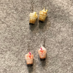 Może przedstawiać: Para kolczyków wiszących z miniaturowymi plastikowymi kubkami z bitą śmietaną i słomką. Jedna para jest brązowa, a druga brzoskwiniowa.