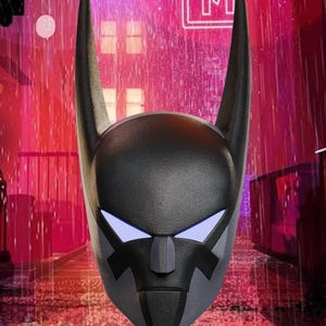 Könnte beinhalten: Eine schwarz-graue Maske mit langen, spitzen Ohren und leuchtend blauen Augen. Die Maske hat eine strukturierte Oberfläche und ein stilisiertes Design, das an einen Superhelden erinnert. Der Hintergrund zeigt eine Stadtlandschaft mit Neonreklamen und Regen.