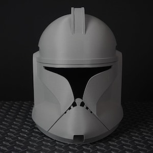 Puede incluir: Un casco gris impreso en 3D, que recuerda al casco de un soldado clon. El casco presenta una parte superior redondeada con una cresta central y una forma de visera distintiva. El diseño incluye detalles angulares en las mejillas y la barbilla, con un interior oscuro.
