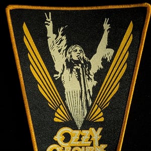 Puede incluir: Parche negro con borde dorado que presenta una ilustración crema de un cantante con los brazos levantados, flanqueado por alas doradas. El texto "OZZY OSBOURNE" está en dorado en la parte inferior. Un parche temático musical.