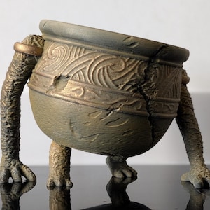 May include: A weathered, bronze-colored pot with arms and legs. The pot has a cracked design and a gold ring around one arm. The arms and legs have a textured, reptilian appearance.