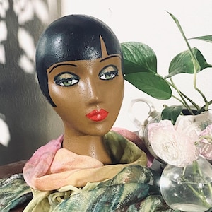 May include: A vintage mannequin head with a painted face, black hair, and red lips. The mannequin is wearing a colorful scarf with pink, green, and yellow hues. A vase with flowers and greenery is in the background.
