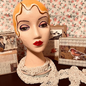 May include: A vintage-style mannequin head with painted features, including red lips and eye makeup, wearing a cream-colored crocheted scarf. Decorative boxes with vintage images and a floral background complete the scene.