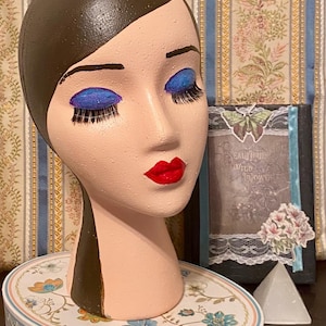 May include: A mannequin head with brown hair and painted features, including blue eyeshadow and red lipstick. The head is on a decorative floral-patterned base with a white bow. A framed picture with the text "BEAUTIFUL WILD FLOWERS" is visible.