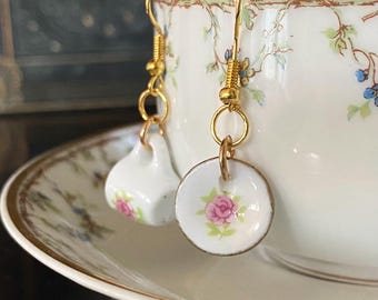 May include: Gold-coloured earrings with miniature white teacups and saucers, adorned with pink rose motifs. The earrings are suspended from gold-toned hooks, presenting a delicate, vintage appearance. The teacups and saucers are decorated with floral patterns.