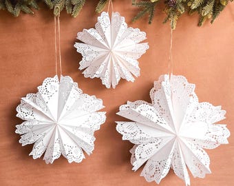 May include: Three white paper star ornaments with intricate lace-like die-cut edges, suspended by thin twine strings. Each star features a multi-pointed, folded design with delicate perforated patterns, hanging against a plain brown background.