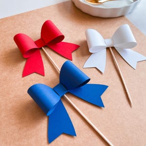 May include: Three paper bows on sticks, one red, one white, and one blue. The bows are arranged on a brown surface.
