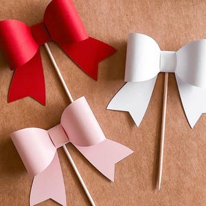 Op de afbeelding: Drie papieren strikken op stokjes, een rode, een witte en een roze. De strikken zijn gemaakt van papier en zijn bevestigd aan houten stokjes. De strikken bevinden zich op een bruine achtergrond.