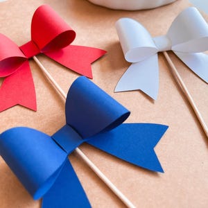 May include: Three paper bows on sticks, red, white, and blue. The bows are made of paper and are attached to wooden sticks. The bows are on a brown background.