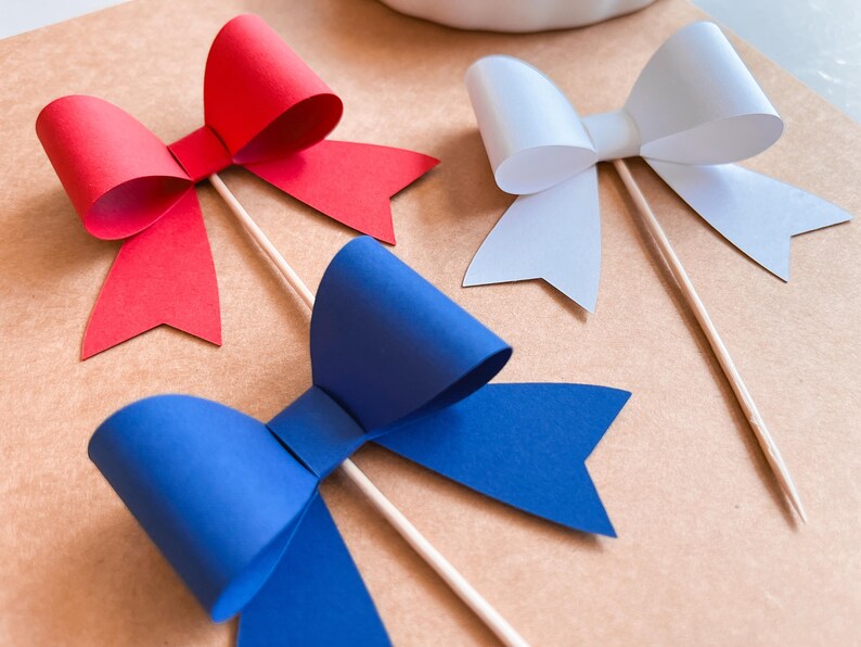 May include: Three paper bows on sticks, red, white, and blue. The bows are made of paper and are attached to wooden sticks. The bows are on a brown background.