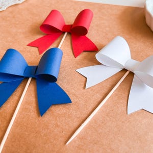 May include: Three paper bows on sticks, one red, one blue, and one white. The bows are made of paper and are attached to wooden sticks.