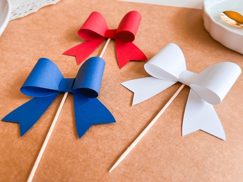 May include: Three paper bows on sticks, one red, one blue, and one white. The bows are made of paper and are attached to wooden sticks.