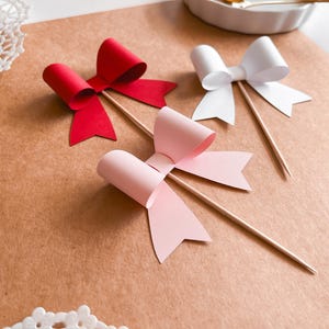 Op de afbeelding: Drie papieren strikken op houten spiesjes. De strikken zijn rood, roze en wit.