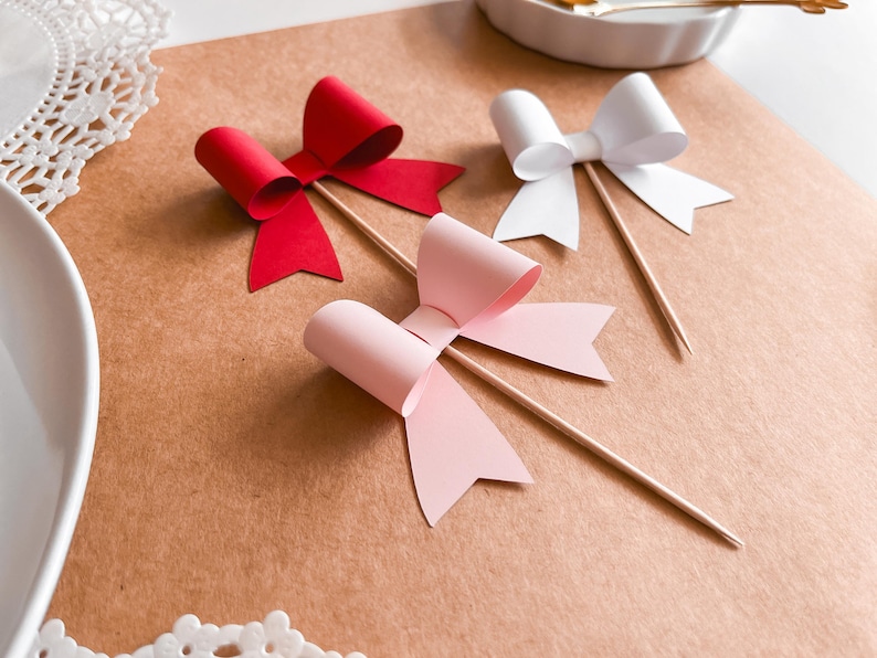 Op de afbeelding: Drie papieren strikken op houten spiesjes. De strikken zijn rood, roze en wit.