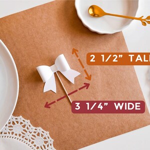May include: White paper bow with a wooden stick, measuring 2 1/2 inches tall and 3 1/4 inches wide. The bow is on a brown surface.
