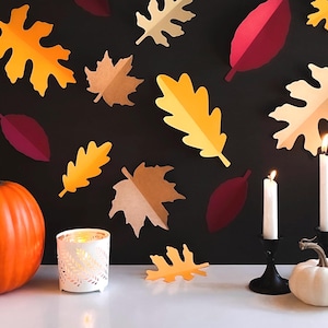 May include: A fall-themed still life with a pumpkin, two white candles in black candle holders, a white candle holder with a cut-out design, and a white pumpkin. The background is black with paper cut-out leaves in shades of orange, yellow, and red.