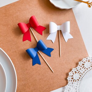 May include: Three paper bows on sticks, red, white, and blue. The bows are arranged on a brown surface.
