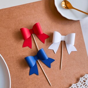 May include: Three paper bows on sticks, one red, one white, and one blue. The bows are on a brown surface.