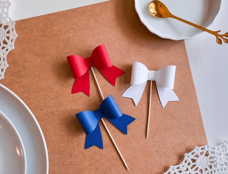 May include: Three paper bows on sticks, one red, one white, and one blue. The bows are on a brown surface.