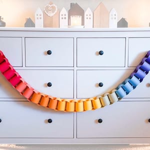 May include: A rainbow-colored paper chain garland featuring interlocking loops in a gradient of red, orange, yellow, green, blue, and purple. The garland is draped across the front of a light gray dresser with six drawers and black knobs.