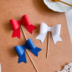 May include: Three paper bows on sticks, one red, one white, and one blue. The bows are arranged on a brown surface.